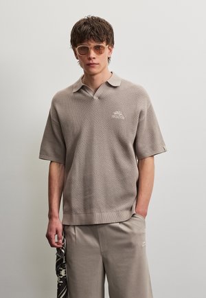 Tânăr purtând un polo bej tricotat cu mâneci scurte, pantaloni asortați, ochelari de soare, ținând un fular cu model pe un fundal simplu, deschis.