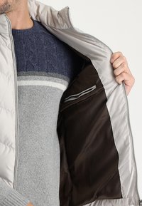 Chaqueta ligera con cremallera en plata, que presenta un exterior acolchado y un forro en marrón oscuro contrastante. Incluye un pequeño bolsillo interior.