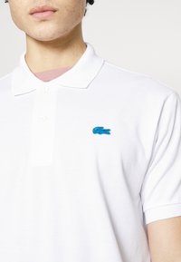 Polo shirt blanc avec un tissu texturé, patte de boutonnage à trois boutons et un logo crocodile brodé bleu sur la poitrine gauche.
