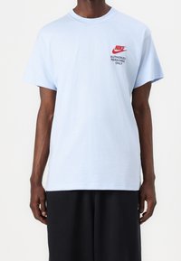 Muž v světle modrém tričku Nike s červeným logem a textem "Pouze pro oprávněný personál" na levé části hrudi, v kombinaci s černými kalhotami.