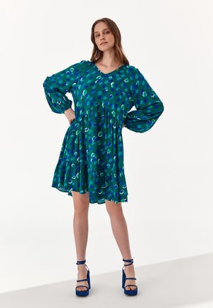 Abito verde con motivi circolari blu e teal, maniche lunghe a palloncino, scollatura a V, orlo arricciato, abbinato a sandali platform blu.