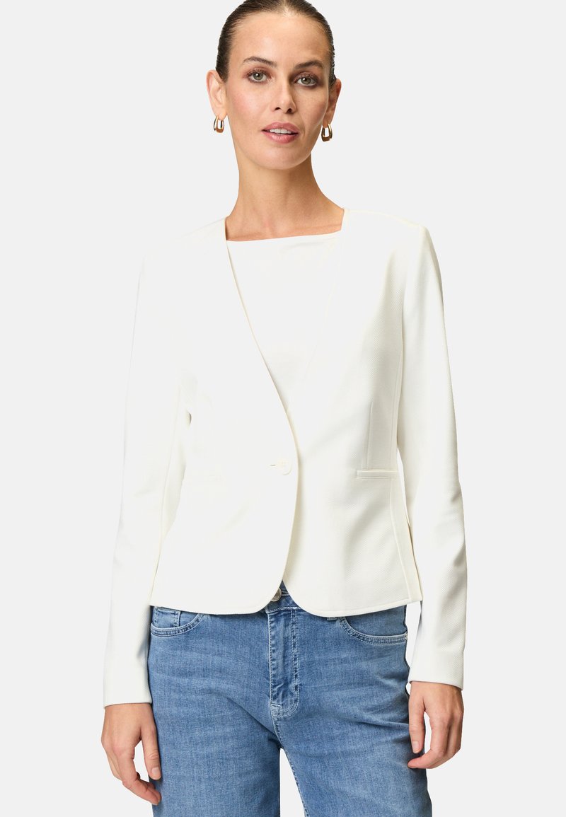 Blazer bianco con una texture liscia, revers a disegno, chiusura con un bottone singolo e due tasche frontali; abbinato a jeans azzurri chiari.
