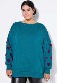 Sudadera de gran tamaño en color teal con mangas largas, presenta acentos en forma de corazón en color burdeos en ambas mangas. Tejido suave, corte holgado, dobladillo acanalado.