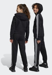 Conjunto de sudadera con capucha y pantalones de chándal negros con rayas blancas en los lados. El material es probablemente una tela suave y texturizada. Incluye una capucha.