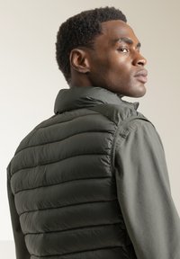 Chaleco tipo puff en verde oscuro, con textura acanalada y cuello alto, superpuesto a una camisa verde clara, mostrando una vista en ángulo desde la parte posterior.