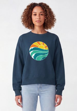 Frau mit lockigem Haar, die einen marineblauen Pullover mit einem bunten Sonnen- und Meereswellenmotiv sowie hellblaue Jeans vor einem einfarbigen Hintergrund trägt.