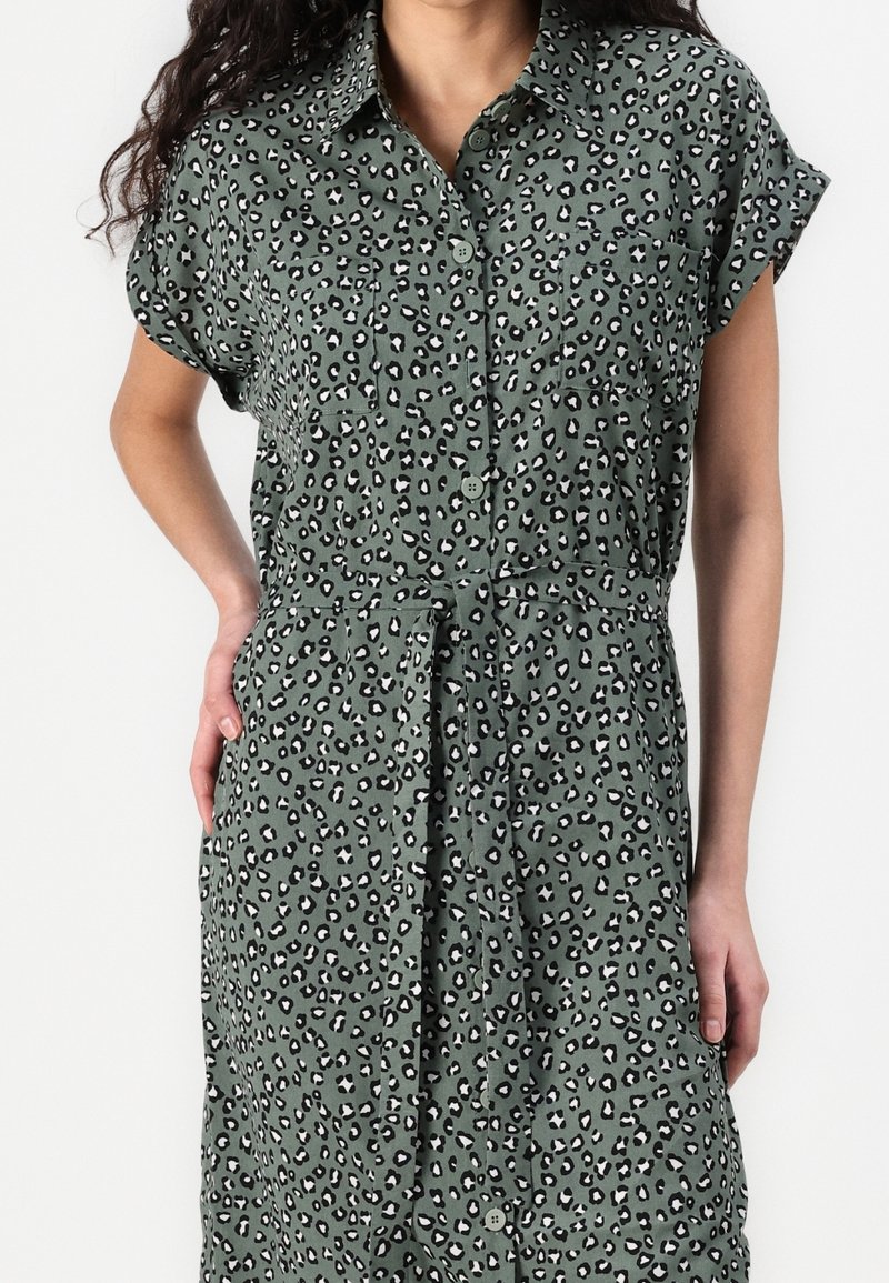 Femme portant une robe boutonnée à manches courtes avec un imprimé léopard vert et noir, ajustée à la taille avec une ceinture en tissu assorti.