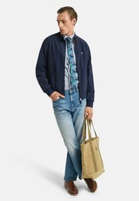 Giubbotto bomber navy, cravatta a fantasia, camicia azzurro chiaro, jeans in denim chiaro, scarpe marroni, borsa beige con manici doppi e logo.