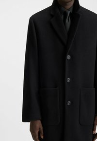 Black wool overcoat featuring a notched lapel, five buttons, and two front pockets. Textured fabric with a smooth finish.