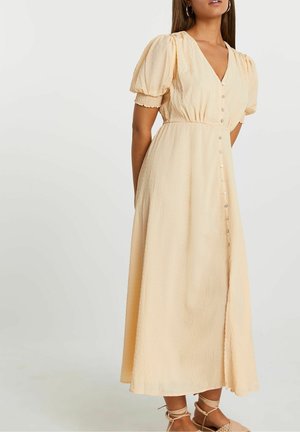 Kvinde iført en lys beige, knappet midi-kjole med puffede korte ærmer og snøresandaler mod en ensfarvet baggrund.