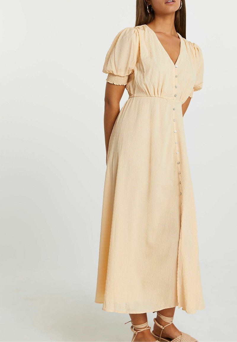 Femme portant une robe midi beige clair boutonnée, avec des manches courtes bouffantes et des sandales à nouer, sur un fond uni.