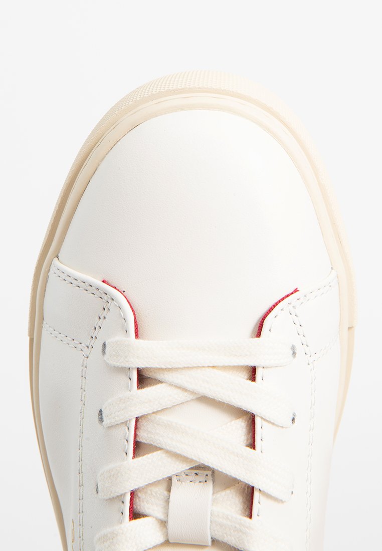 Vita läder sneaker med rund tå, beige gummisula och vita snören. Har röd accent nära öglorna och synliga sömnadsdetaljer.