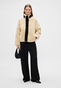 Κρεμ γούνα σακάκι με υψηλό γιακά και μαύρο μπροστινό πάνε�λ, συνδυασμένο με μαύρο παντελόνι με φαρδιές ποδιές. Το μοντέλο κρατά μια μικρή μαύρη τσάντα.