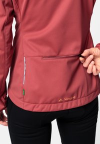 Rød softshell-jakke med en glidelåslomme bak, kontrasterende lyse sømmer, logo i oransje, og en glatt tekstur. Svarte bukser synlige.