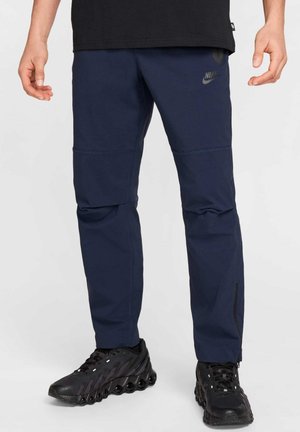 Homme portant un pantalon Nike bleu marine avec coutures avant et des chaussures de sport noires avec une semelle amortissante unique, debout sur un fond clair.