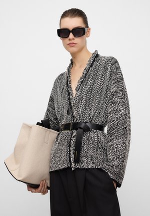 Frau mit großen schwarzen Sonnenbrillen, schwarz-weiß strukturiertem Cardigan, am Taillenbereich mit Gürtel, hält eine große beigen Tragetasche vor weißem Hintergrund.