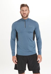 Langärmliges Sportshirt in Blau mit einem halben Reißverschluss an der Vorderseite, schwarzen seitlichen Einsätzen und einem glatten, feuchtigkeitsableitenden Stoff.