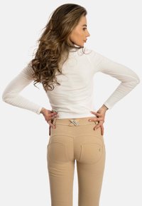 Beige korkeavyötäröiset housut, joissa on tiukka muotoilu, näkyvät sauma-yksityiskohdat ja koristeelliset tikkaus. Vyötäröllä oleva etiketti osoittaa brändin.