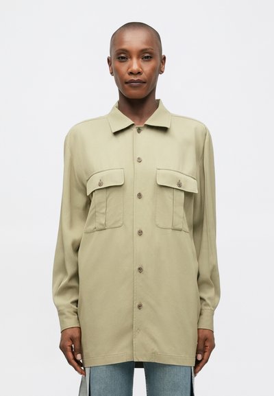 Camicia button-up verde oliva con due tasche sul petto, maniche lunghe e vestibilità rilassata. Il tessuto appare liscio con una leggera lucentezza.