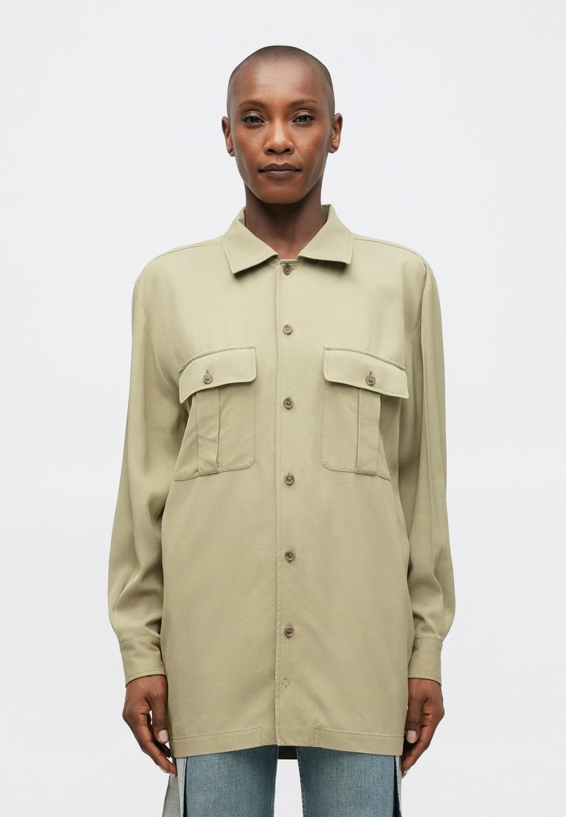 Alyvinės žalios spalvos marškiniai su sagomis, dviem krūtinės kišenėmis, ilgomis rankovėmis ir laisvu siluetu. Audinys atrodo lygus su švelniu blizgesiu.