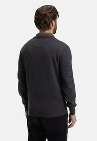 Homme avec les cheveux bruns courts portant un pull gris foncé par-dessus une chemise verte à col, vu de dos sur un fond uni.