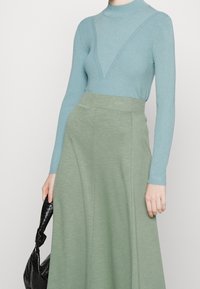 Hellblau gestreift, langärmeliges Oberteil mit V-Ausschnitt, kombiniert mit einem fließenden grünen Midi-Rock. Schwarze, strukturierte Handtasche in der Hand.