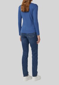 Frau, die von hinten schaut, trägt ein blaues Langarmoberteil, dunkelblaue Jeans und weiße Sneaker vor einem schlichten grauen Hintergrund.
