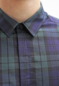 Rutigt skjorta med knäppkrage. Tyget har mörkgröna, blå och svarta rutor. Slät textur med synlig sömnad och detalj med knappar.