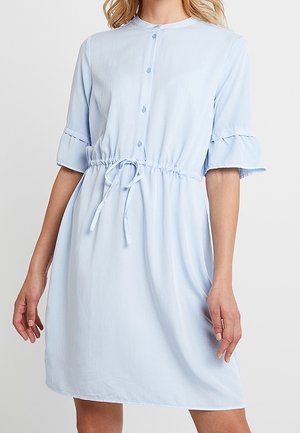 Vaalean sininen, polveen ulottuva mekko, jossa on lyhyet röyhelöhihoja, napitus edessä ja säädettävä vyötärönauha, jota kantaa seisova henkilö.