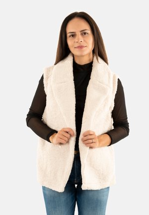 Donna con lunghi capelli scuri, che indossa un top nero a maniche lunghe, jeans blu e un gilet bianco senza maniche, mentre tiene i bordi del gilet.