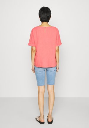 Person mit kurzen dunklen Haaren steht mit dem Rücken zur Kamera, trägt ein korallenfarbenes kurzärmeliges Oberteil, hellblaue knielange Jeansshorts und Sandalen.