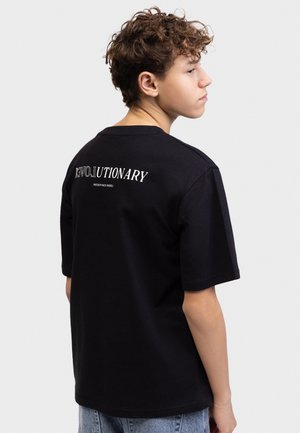 Tenåring med krøllete hår iført svart T-skjorte med teksten "REVOLUTIONARY" på baksiden, stående mot en ensfarget lys bakgrunn.