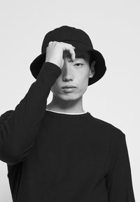 Chapeau seau noir et chemise à manches longues noire avec un bord blanc. La personne est partiellement obscurcie, touchant son front avec une main.