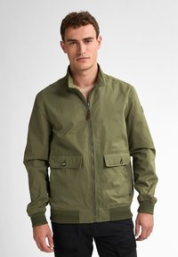 Jachetă bomber de culoare verde măsliniu, cu guler înalt, fermoar frontal și două buzunare cu clapă și nasturi. Include manșete și tiv din tricot.