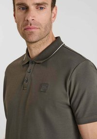 Polo de color verde oscuro con cuello, tapeta de tres botones y acentos blancos contrastantes en el cuello. Presenta un parche de logo en el pecho.
