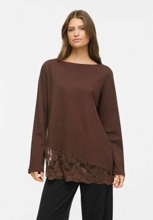 Femme portant un haut marron à manches longues avec un détail en dentelle diagonale à l'ourlet, associé à un pantalon noir, debout devant un fond uni.