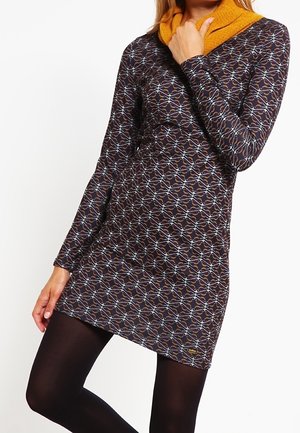 Kobieta ubrana w długą sukienkę z wzorem, z kołnierzem w kolorze musztardowej żółci, w połączeniu z czarnymi, kryjącymi rajstopami, stojąca na tle białego tła.