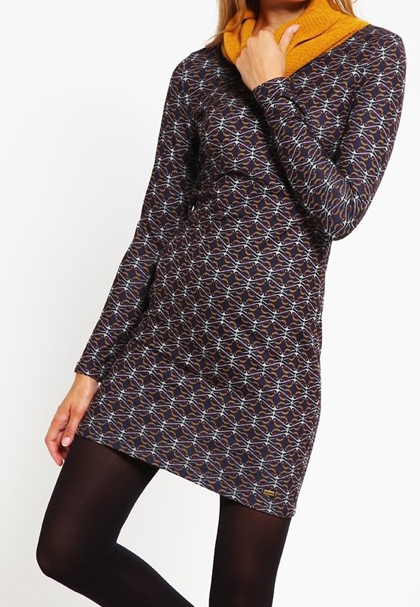 Femme portant une robe à manches longues à motifs avec un col jaune moutarde, associée à des collants opaques noirs, debout devant un fond blanc.