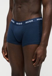 Buste masculin portant un boxer bleu marine avec le logo "HUGO" sur la ceinture, debout devant un fond clair uni.