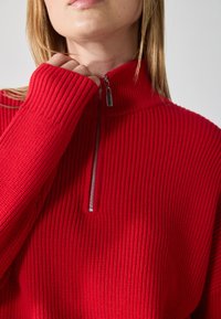 Červený žebrovaný svetr na zip s vysokým límcem a stříbrným zipem, který vystavuje strukturovaný pletený vzor. Detailní záběr na oblast krku.