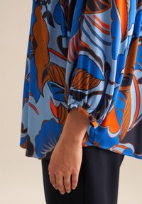 Blusa larga e a manica lunga in tessuto blu con vivaci motivi floreali arancioni e marroni. Include dettaglio dei bottoni ai polsini.