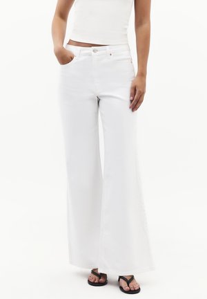Kvinde iført hvide brede jeans, hvid kort top og sorte thongsandaler, står med den ene hånd i lommen.