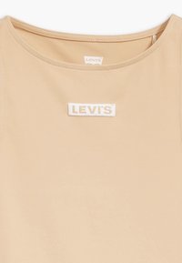Top sin mangas beige con un escote redondo y una etiqueta blanca de Levi's incrustada en la parte frontal. Textura suave y diseño minimalista.