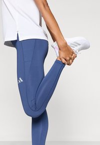 Modré športové legíny z pružného materiálu, s priliehavým strihom, kontrastným prešitím a logom Adidas na stehne. Biela dekorácia na topánke.