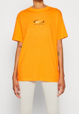 Person iført en skrigorange Nike Sportswear t-shirt og lyse bukser, stående mod en ensfarvet hvid baggrund.