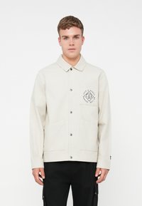 Giacca beige chiaro con colletto, chiusura frontale con bottoni e due tasche frontali. Presenta una patch con il logo sul petto e una texture sottile.