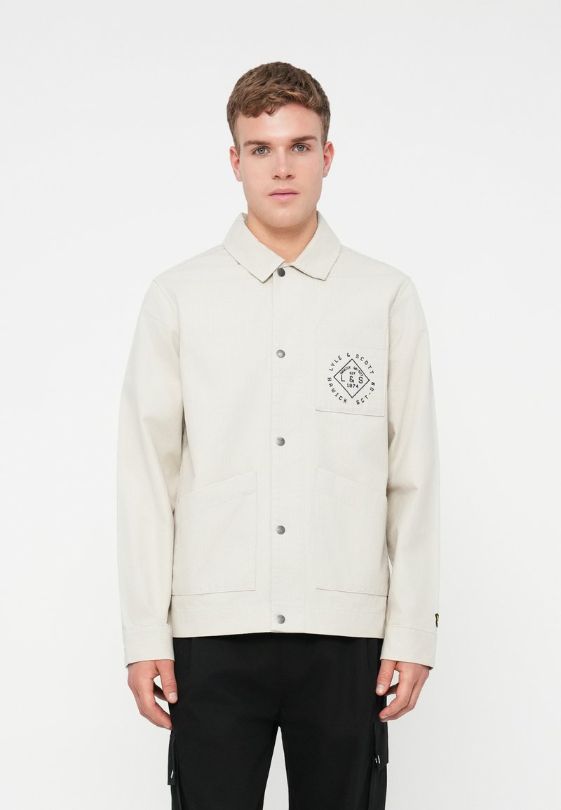 Giacca beige chiaro con colletto, chiusura frontale con bottoni e due tasche frontali. Presenta una patch con il logo sul petto e una texture sottile.