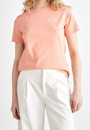 Kobieta ubrana w brzoskwiniowy t-shirt z krótkim rękawem z napisem "SAINT 1949 N.H.C" oraz białe plisowane spodnie z wysokim stanem na białym tle.