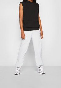 Black sleeveless hooded sweatshirt with a front pocket paired with light gray sweatpants. White athletic shoes complete the outfit.