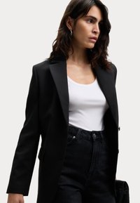 Černý blazer s klopami, jedno tlačítko, a bočními kapsami, nošený přes bílé tílko a tmavé džíny.